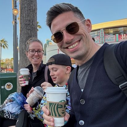 A picture of Aaron Aiken, smiling directly at the camera, on a bench with his family enjoying beverages from Starbucks.